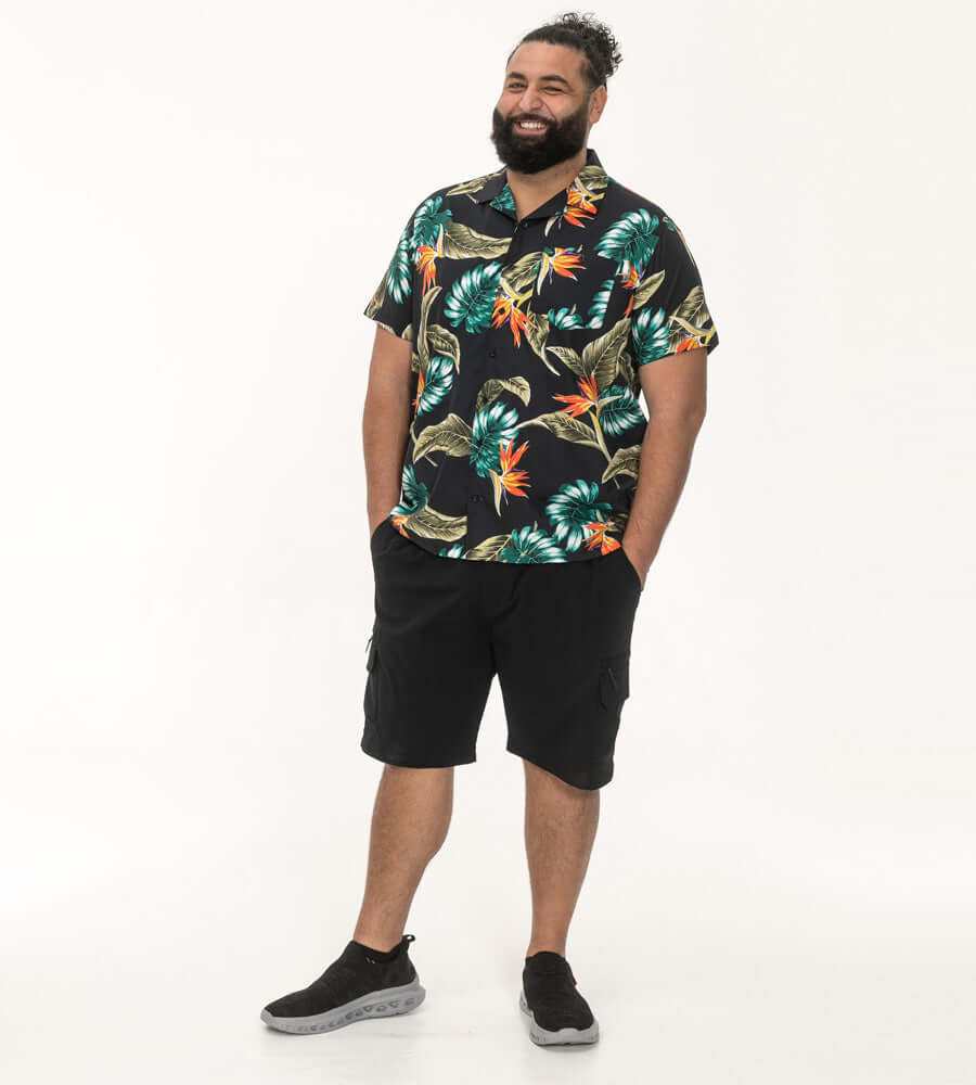 Model in een zwart Hawaiian polo met tropische print, gecombineerd met donkere shorts en sneakers.