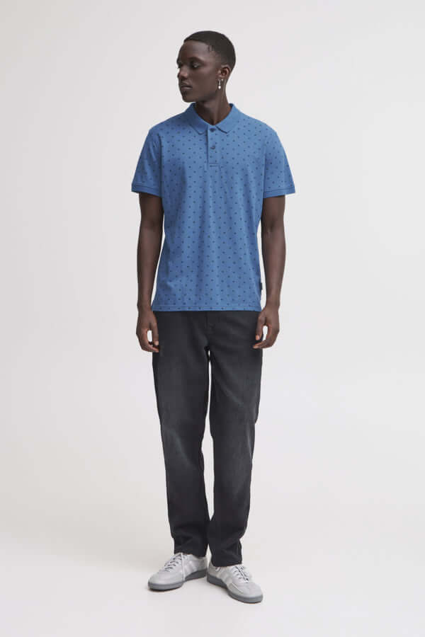 Man draagt een blauwe polo met opdruk, gecombineerd met zwarte broek en sneakers, perfect voor casual outfits.