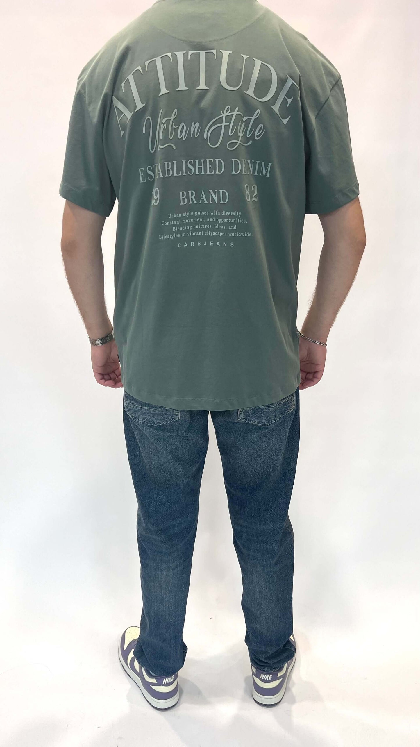 Man achteraan in groene T-shirt met 'Attitude' op print en blauwe jeans.