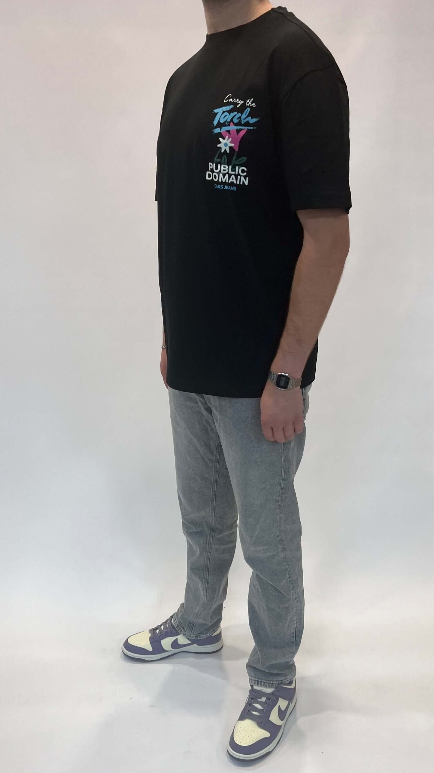 Man in zwart T-shirt met kleurrijke opdruk, grijze jeans en sneakers, staand tegen witte achtergrond.