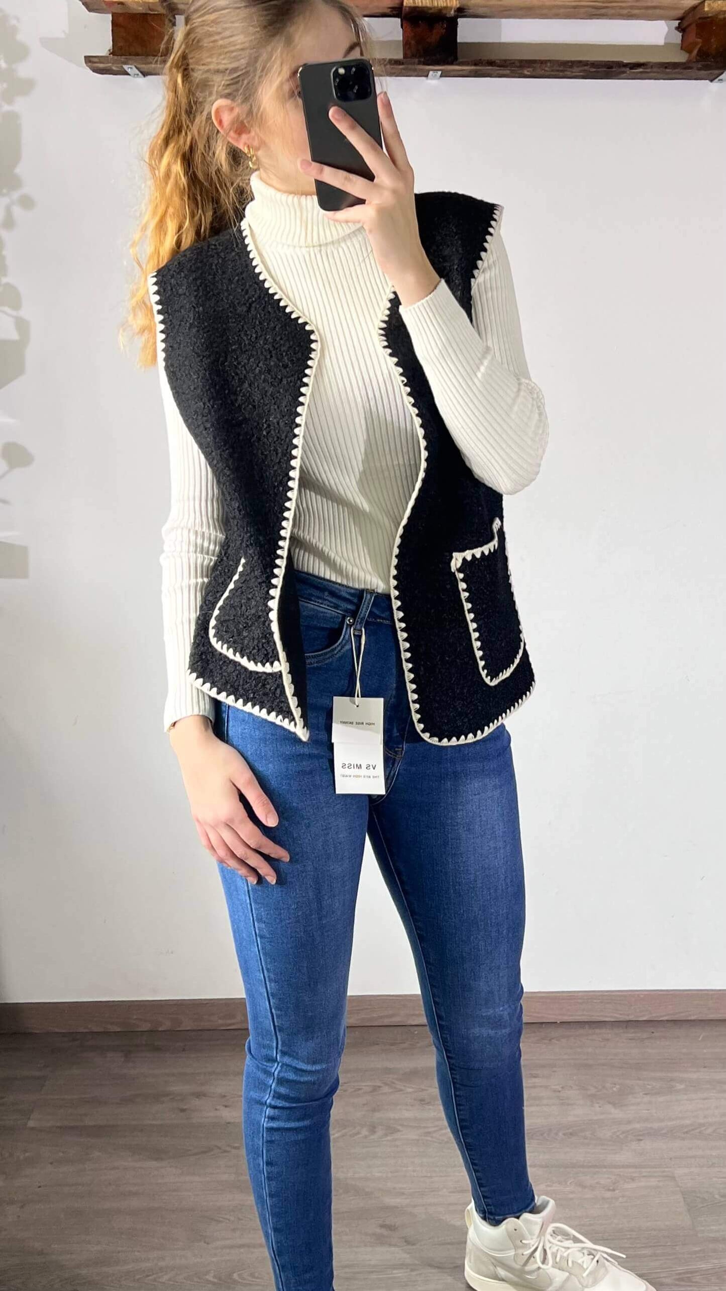 Vrouw in zwart gilet met witte details, gecombineerd met een witte coltrui en jeans.